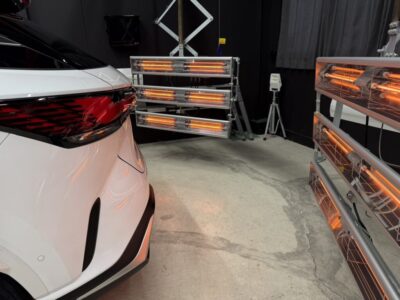 White car seen from the rear in a workshop with orange infrared heat lamps on racks around it and at the right edge of the image.