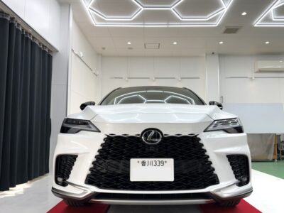 Front view of a white Lexus sedan in a bright showroom with geometric ceiling lights and a red carpet.