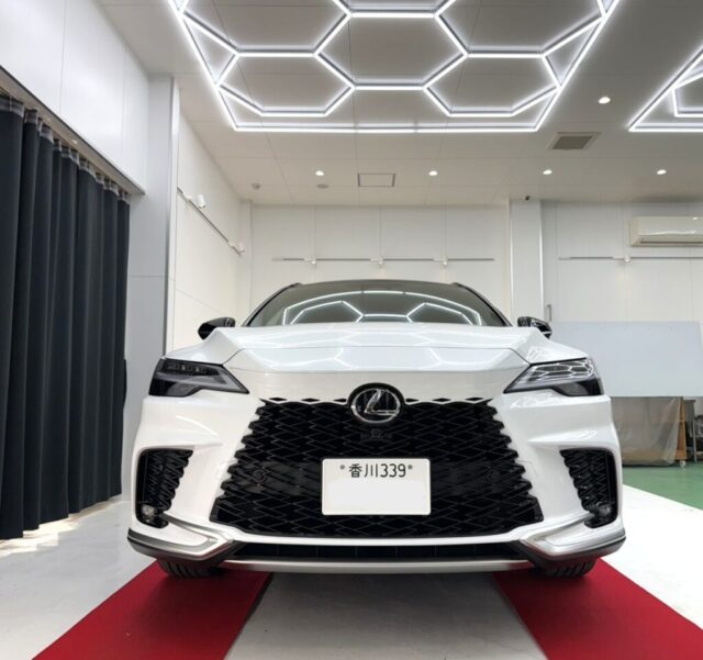 Front view of a white Lexus sedan in a bright showroom with geometric ceiling lights and a red carpet.
