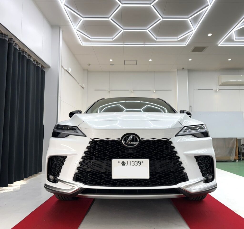 Front view of a white Lexus sedan in a bright showroom with geometric ceiling lights and a red carpet.