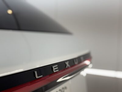 Close-up of a white car's trunk showing the Lexus badge along a dark chrome trim with a red accent line.