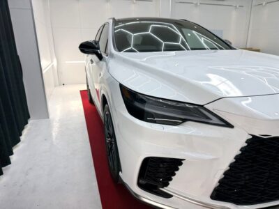 White luxury sedan on display in a bright showroom, viewed from the front-right with a red carpet underneath.