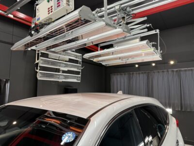 White car in a dark studio with large overhead infrared lamps and industrial equipment suspended from the ceiling above it.
