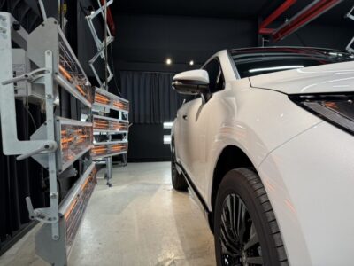 White car in a modern workshop, with orange-tube heating lamps along the left wall and equipment nearby, viewed from the front-right side of the vehicle.