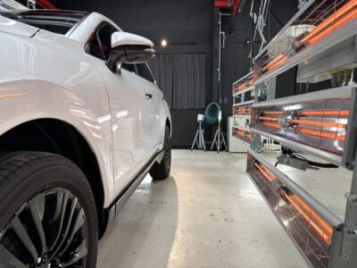 Side view of a white SUV inside an interior testing chamber, with orange infrared heater panels on the right and equipment in the background.