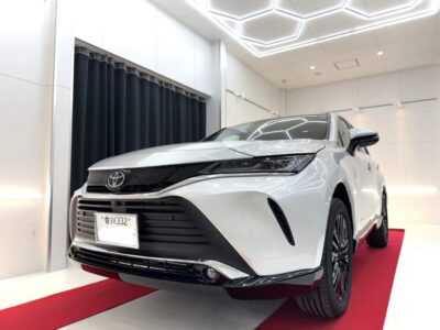 Front 3/4 view of a white Toyota SUV on a red carpet in a bright, modern showroom with geometric ceiling lights.