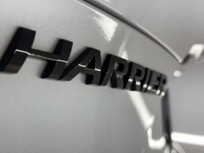 Close-up of a car's rear badge spelling 'HARRIER' on metallic gray paint, with reflections on the surface.