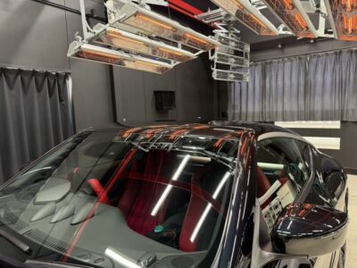Black car with red interior parked inside a lab-like room, with ceiling-mounted infrared heaters and equipment above and gray curtains in the background.