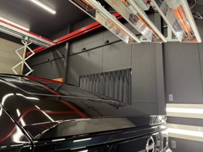 Black Nissan SUV in a garage-like workshop with bright ceiling heaters and industrial piping above the car surface reflecting light.