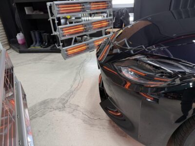 Automotive shop scene with a glossy black car and orange infrared heater panels nearby for curing paint or coatings.