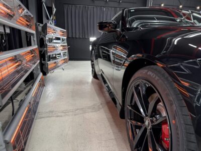 Black sports car inside a showroom with orange infrared heaters along the wall at left edge