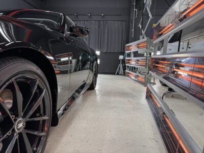 Black sports car in a studio with orange infrared heat lamps along the right wall.