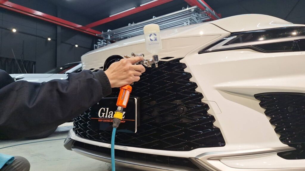 Worker uses a spray gun to apply coating on the front grille of a white car in a detailing shop.