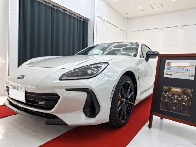 White sports car on a red carpet in a bright showroom, shown from the front-left, with black alloy wheels and sleek headlights.