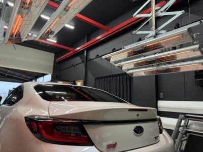 White Subaru WRX STI rear view inside a modern auto workshop with orange infrared heaters overhead and dark wall panels nearby.