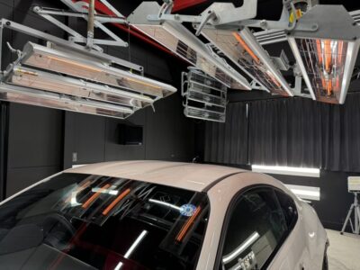 White car in a dark showroom or lab surrounded by ceiling-mounted infrared heaters for paint curing or testing operations.