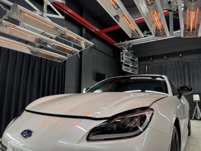 White sports car photographed from the front-left in a testing studio with overhead infrared heaters and dark curtains behind.