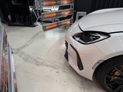 White car front-right corner in a showroom, with orange infrared heaters visible in the background and a glossy black wheel nearby.