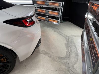 White car in a garage with orange infrared heater lamps nearby and a cracked concrete floor view from the rear-left side of the car's tail area