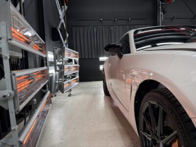 White sports car parked in a workshop with orange infrared heaters lining metal racks on the left and a dark backdrop in the background.