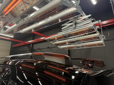 Black SUV in a workshop with roof rails and a ceiling-mounted rack of infrared heating lamps above it