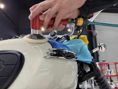 Person buffing a cream-and-blue Triumph motorcycle fuel tank with a red polisher hand tool in a workshop
