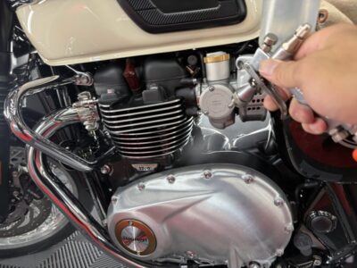 Close-up of a Triumph motorcycle engine with chrome exhaust pipes and cooling fins; a hand holds a tool near the carburetor. From midsection of bike.