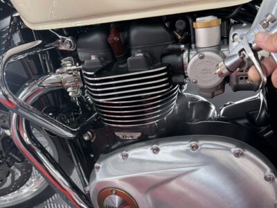 Close-up of a Triumph motorcycle engine showing cooling fins, chrome exhaust, and a beige fuel tank; a hand is operating the carburetor/fuel tap.