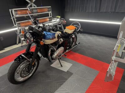 Blue and cream Triumph motorcycle parked on a red-and-gray patterned workshop floor with infrared heaters in the background.