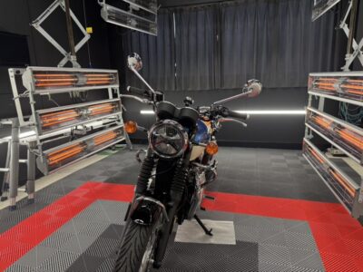 Motorcycle in a test lab with orange infrared heaters on both sides and a checkered red-gray floor behind it