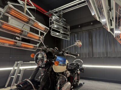 Motorcycle in a high-tech test chamber with orange infrared heaters above and metallic racks around the bike, Triumph logo visible on the tank