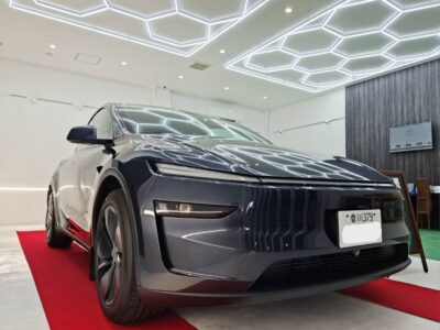 Front three-quarter view of a sleek gray electric car on a red carpet in a bright showroom with hexagonal LED ceiling lights reflected on the hood.