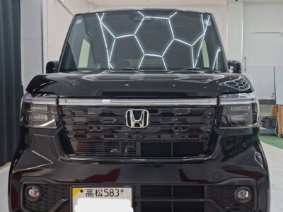 Front view of a black Honda van in a showroom, chrome grille with Honda emblem and reflective hexagonal ceiling lights above.