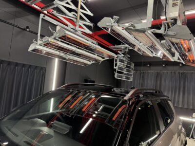 Black SUV in a dark garage, with ceiling-mounted metal rack system and orange infrared heater bars above the vehicle.