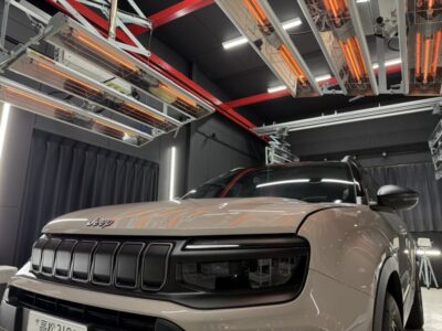Beige Jeep SUV in a showroom or lab with overhead infrared heaters and industrial lighting above, reflecting on the car's hood and window.