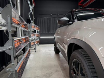 White SUV parked indoors beside orange infrared heating panels mounted on the left wall of a workshop.