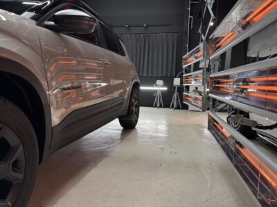 Beige SUV in a testing lab with orange infrared heaters on racks to the right and a dark backdrop ahead.