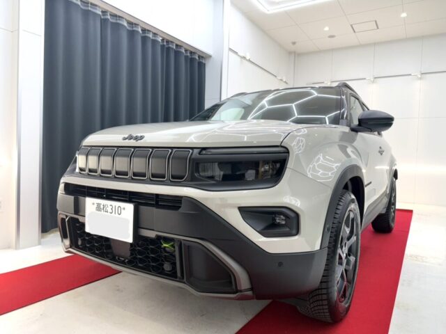 Gray Jeep SUV on a red carpet inside a showroom, shown from the front-right angle, headlights on.