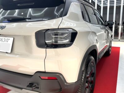 White Jeep SUV on a red showroom carpet, shown from the rear-right side with prominent taillight and hitch, indoor dealership setting.