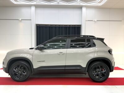 Side view of a gray Jeep Avenger compact SUV with black roof rails and black wheels, displayed indoors on a red carpet.