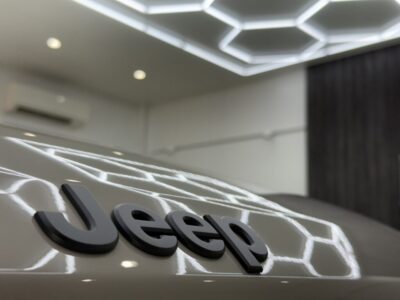 Close-up of a white car with a black Jeep emblem on the hood in a showroom setting with hexagonal ceiling lights.