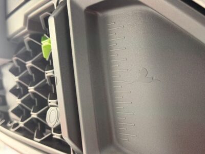 Close-up of a car interior showing a textured plastic panel with vertical measurement-like marks and a small embossed logo, beside a honeycomb vent and a green component in the background.