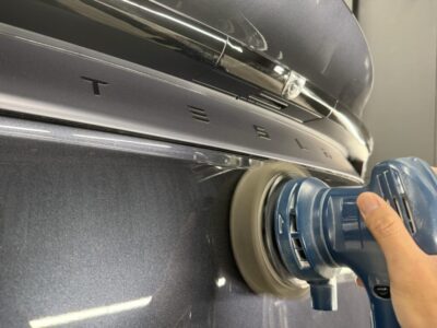 Polisher being used on the rear of a dark blue Tesla, buffing the TESLA badge on the trunk area.