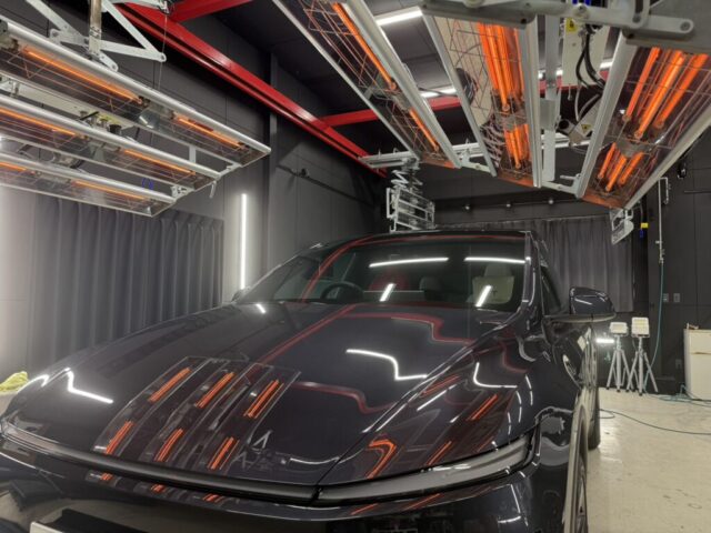 Black SUV in a lab-like testing chamber with orange infrared heater panels overhead and gray curtains nearby.