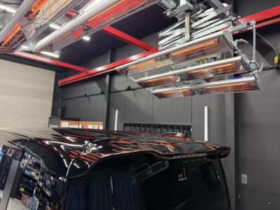 Indoor auto workshop with a glossy black van and orange infrared curing lamps overhead on red metal rails