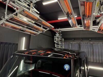 Black SUV inside a lab or test chamber with overhead infrared heating lamps and testing equipment above it.