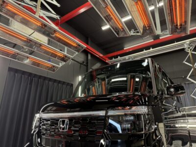 Shiny black Honda minivan inside a high-tech testing facility with overhead infrared heaters above.