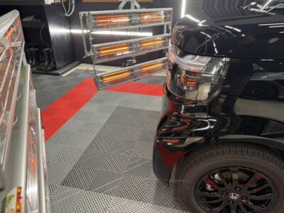 Front-right of a glossy black SUV in a detailing shop, with orange infrared heater racks and a red stripe on a gray checkered floor in view.