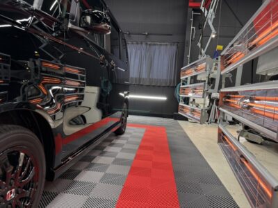 Black van in a professional auto-detailing/shop with orange infrared lamps along the right and a red-gray tiled floor.