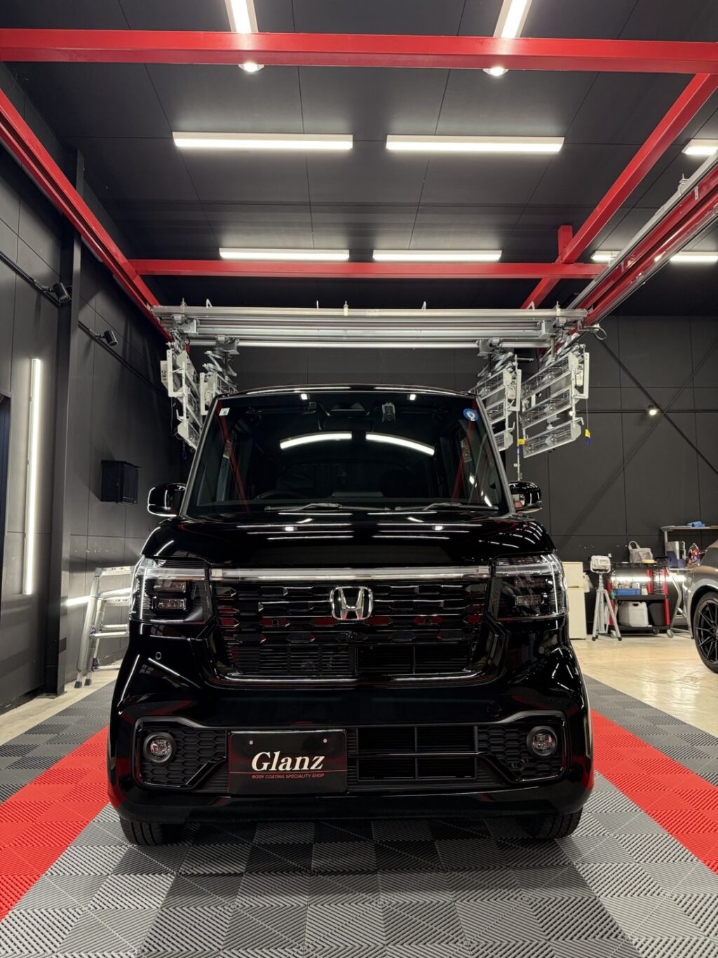Front view of a shiny black Honda van parked inside a modern auto detailing shop with red steel beams overhead and bright ceiling lights.